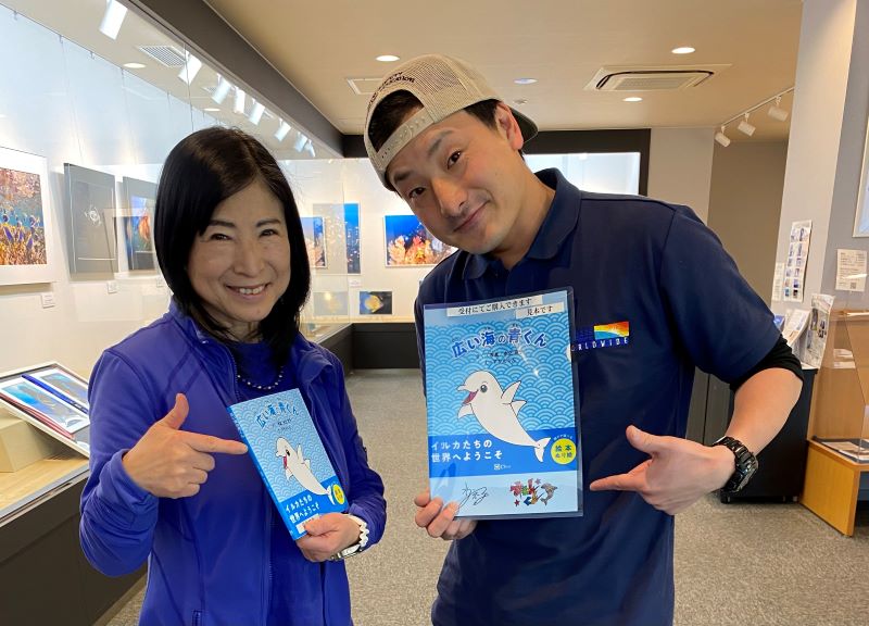 写真展会場の水之京子さんと、「ずかんくん」こと田中一秀さん。手にしているのが２人のコラボ絵本『広い海の青くん』